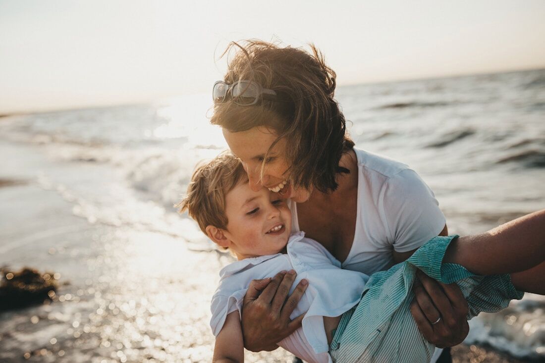 A mother lifting her laughing son at the shoreline in soft evening light