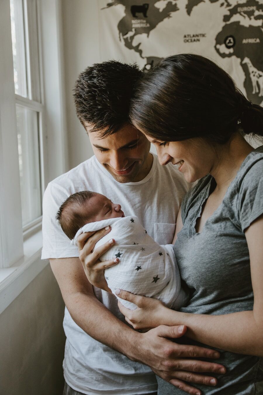 Two parents by a bright window holding their newborn, wrapped in white linen